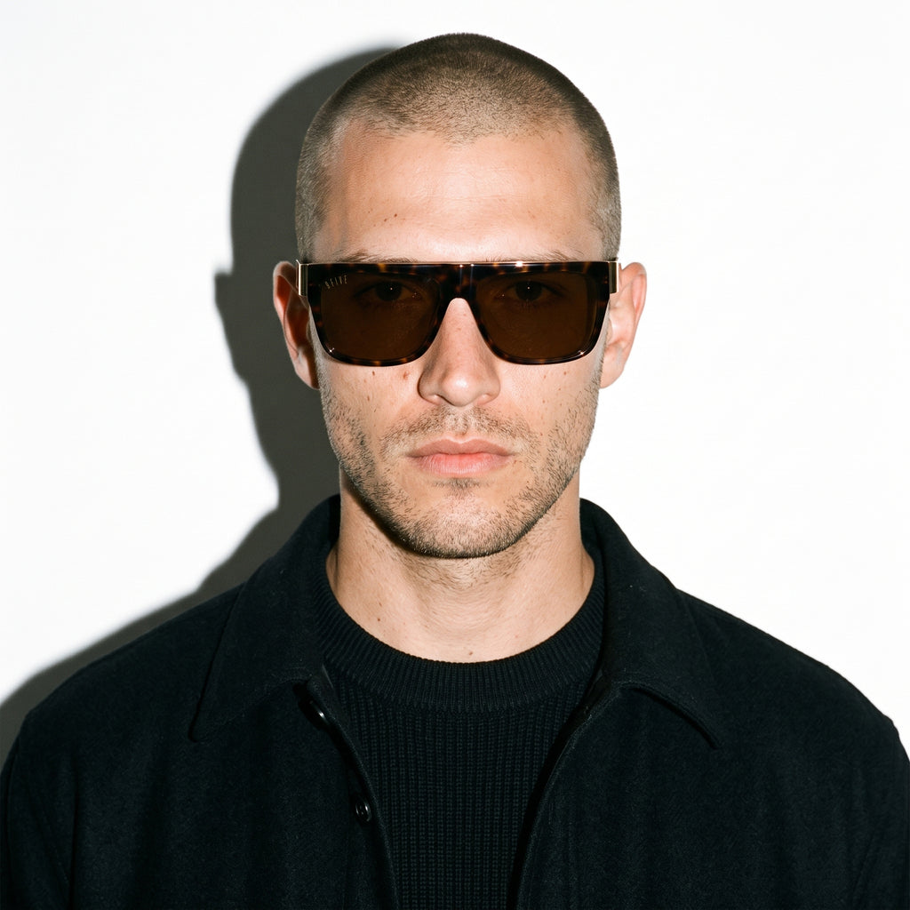 A man with a shaved head wearing dark brown square sunglasses and a black zip-up jacket over a black ribbed sweater, photographed against a white background with a strong shadow.
[g:brown s:all o:3]