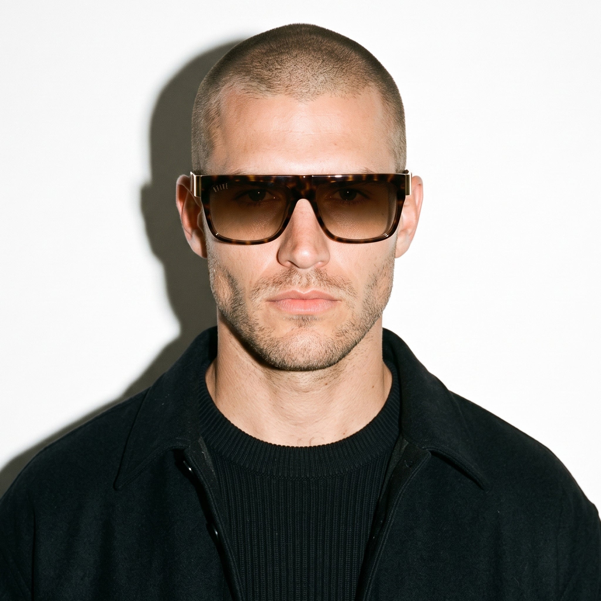 A man with a buzz cut wearing large square tortoiseshell sunglasses and a black zip-up jacket over a black ribbed sweater, photographed against a white background with a hard shadow.
[g:brown-gradient s:all o:3]