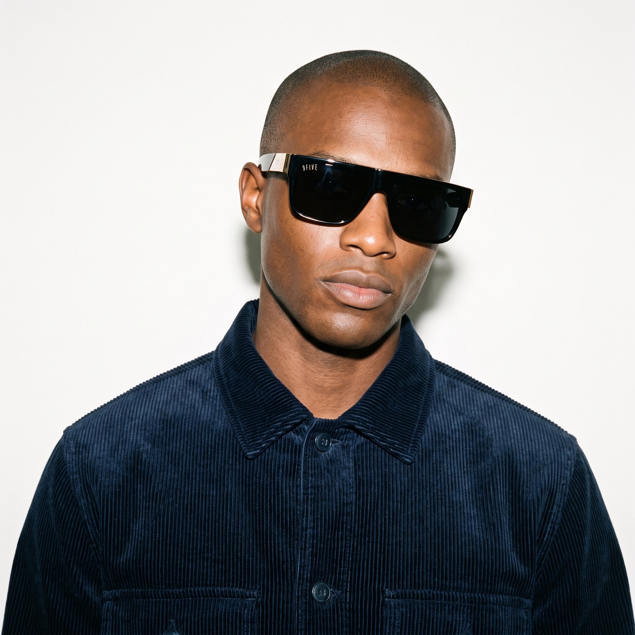 Man wearing dark sunglasses and a navy blue corduroy shirt against a white background [g:black s:all o:3]