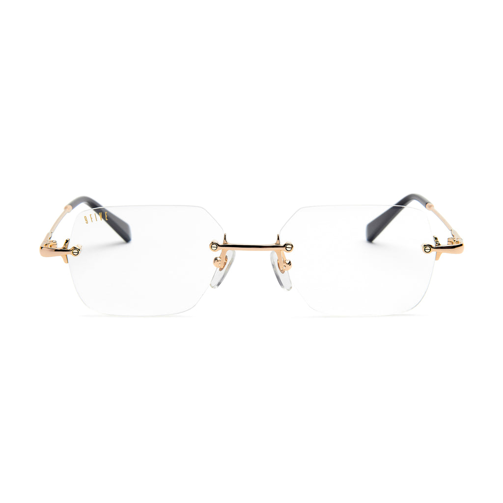 Rimless eyeglasses with clear lenses, gold metal bridge and hinges, and dark gray temple arms on a white background.