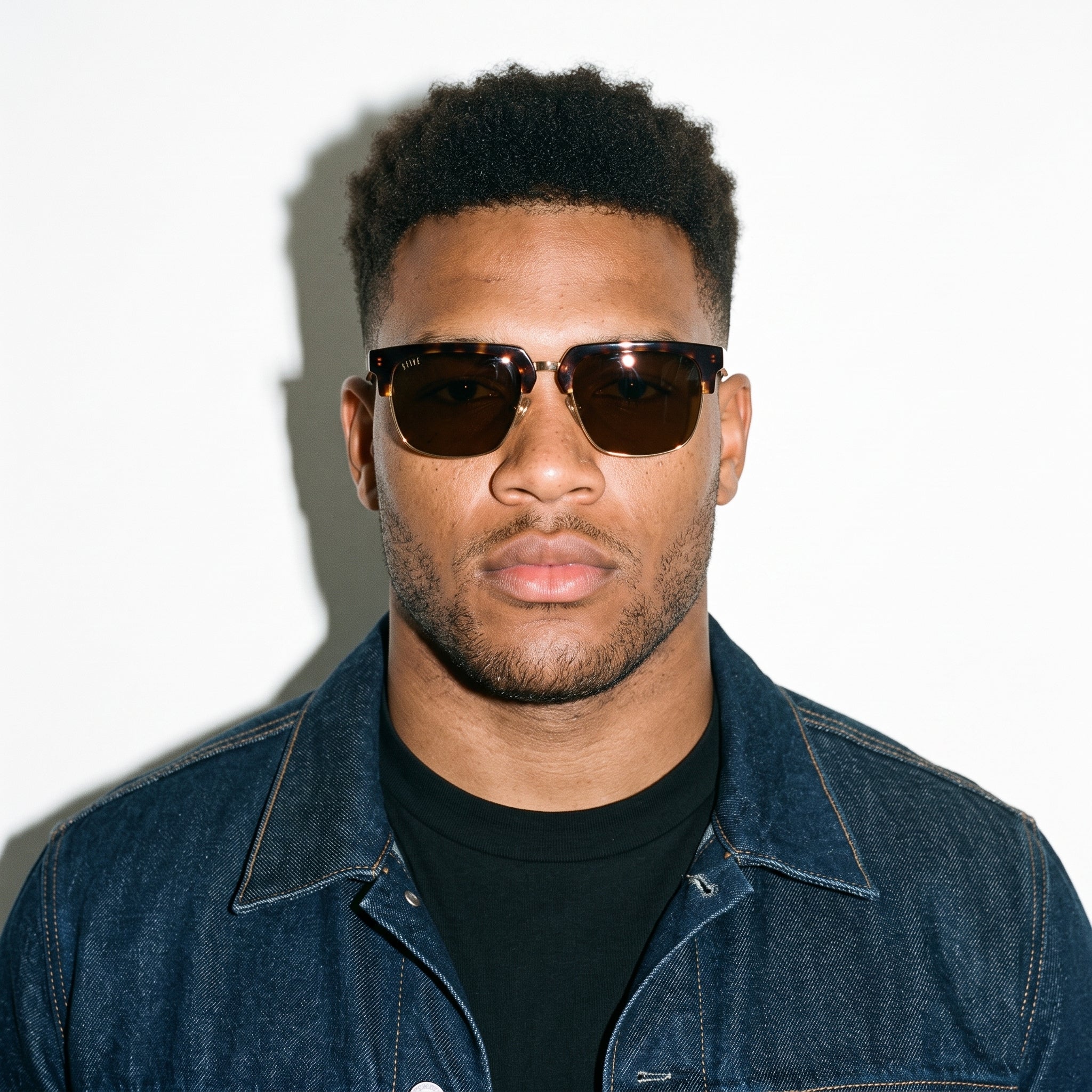 A man wearing dark brown aviator-style sunglasses with tortoiseshell frames, a black crewneck t-shirt, and a blue denim jacket, photographed against a white background with a hard shadow. [g:brown s:all o:3]