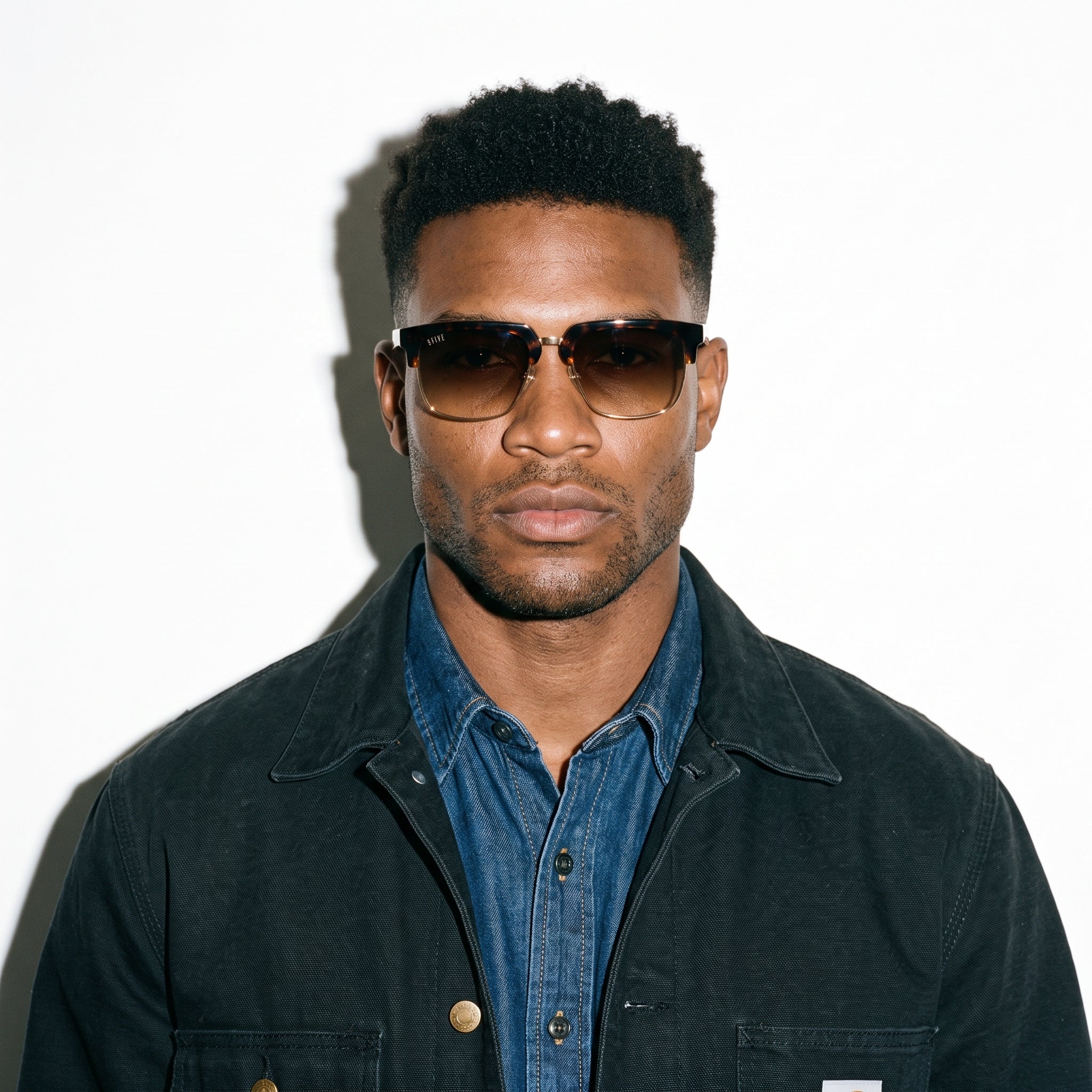 A man wearing dark brown aviator-style sunglasses, a black jacket, and a blue denim shirt, photographed against a plain white background with a strong shadow.
[g:brown-gradient s:all o:3]