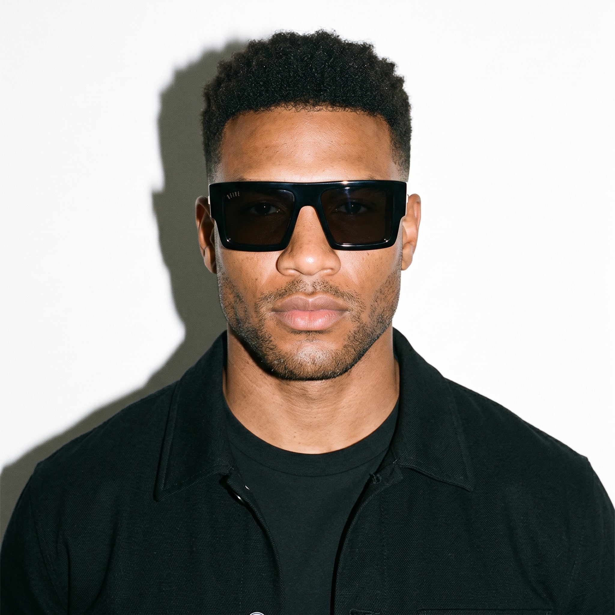A man wearing black square sunglasses and a black collared shirt, photographed against a plain white background with a strong shadow. [g:blacks:all o:3]