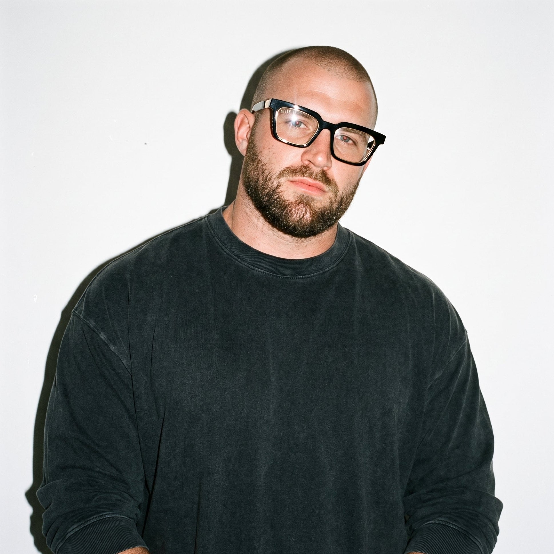 A man with a shaved head, beard, and black-rimmed glasses wearing a dark charcoal long-sleeve crewneck shirt, photographed against a plain white background. [g:clear s:all o:3]
