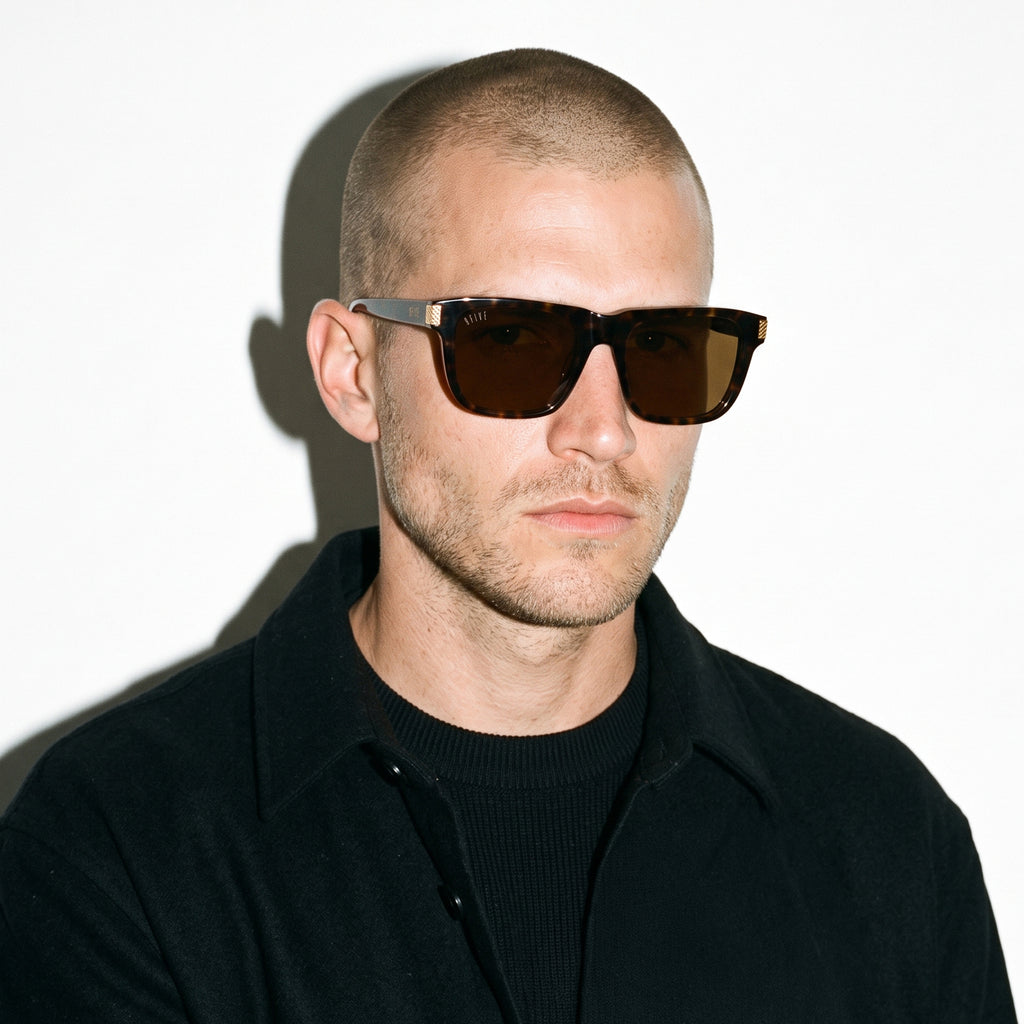 A man with a shaved head wearing dark brown rectangular sunglasses and a black collared shirt, photographed against a plain white background with a strong shadow.[g:brown s:all o:3]