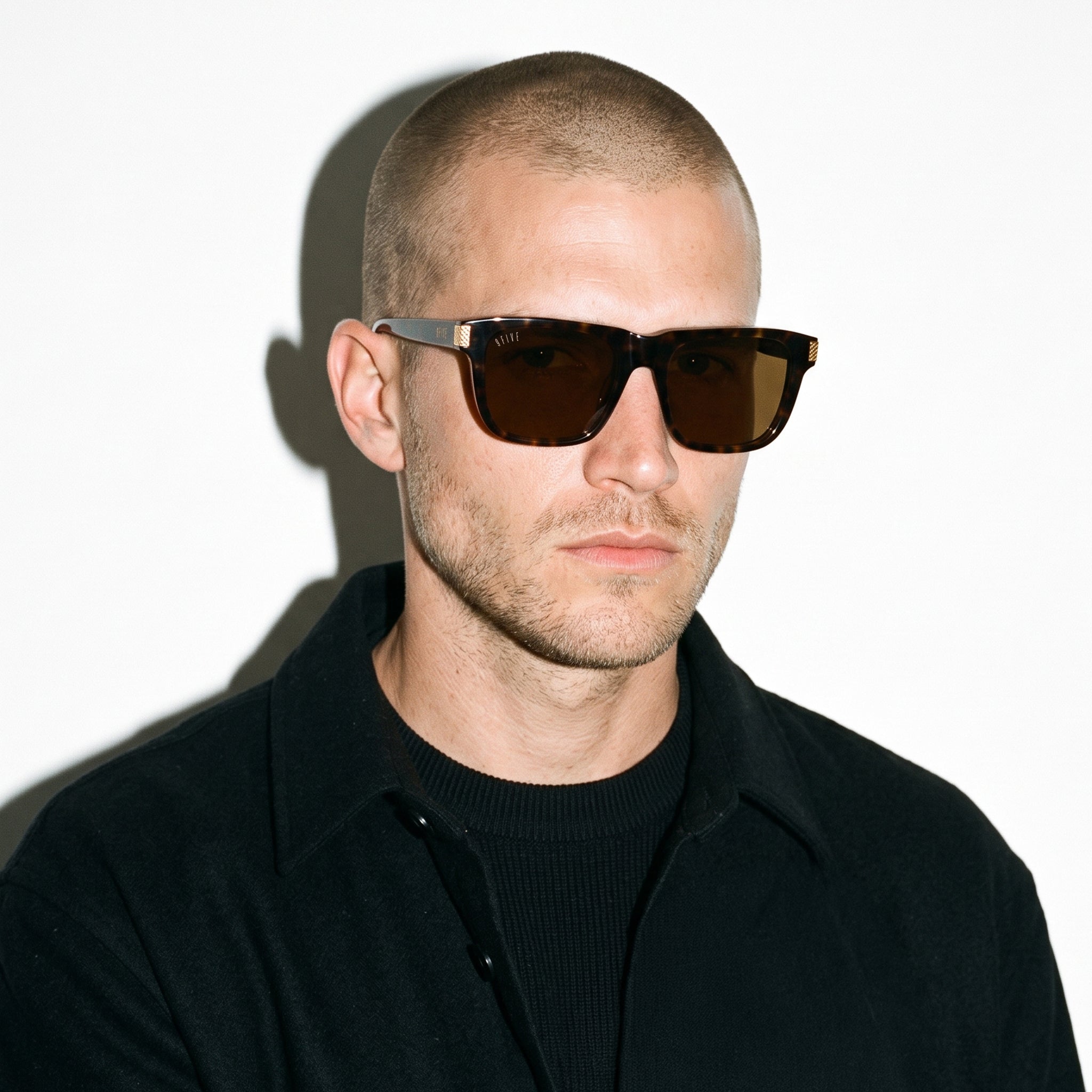 A man with a shaved head wearing dark brown rectangular sunglasses and a black collared shirt, photographed against a plain white background with a strong shadow.[g:brown s:all o:3]