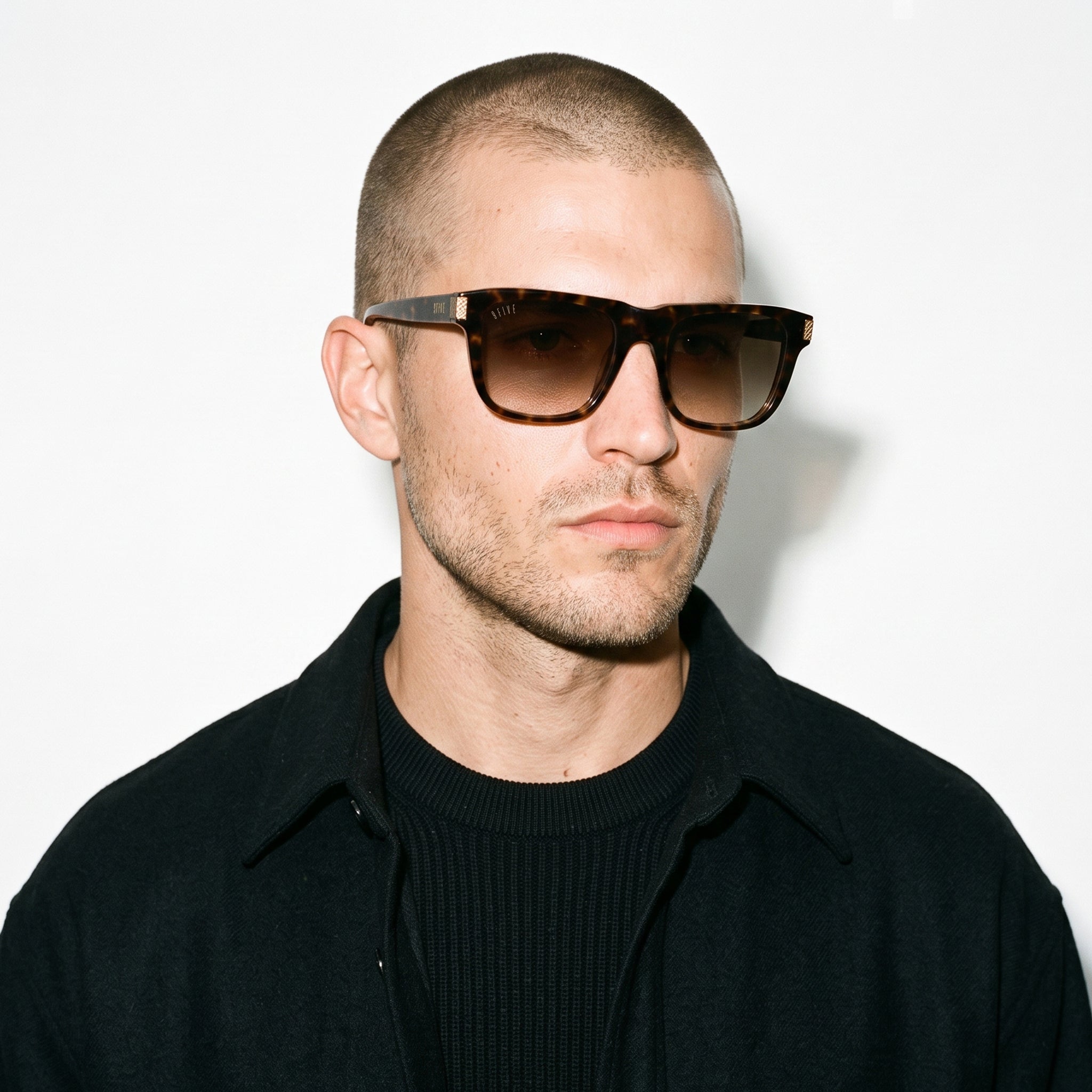 A man with a buzz cut wearing tortoiseshell sunglasses with dark lenses and a black collared shirt, photographed against a plain white background.[g:brown-gradient s:all o:3]