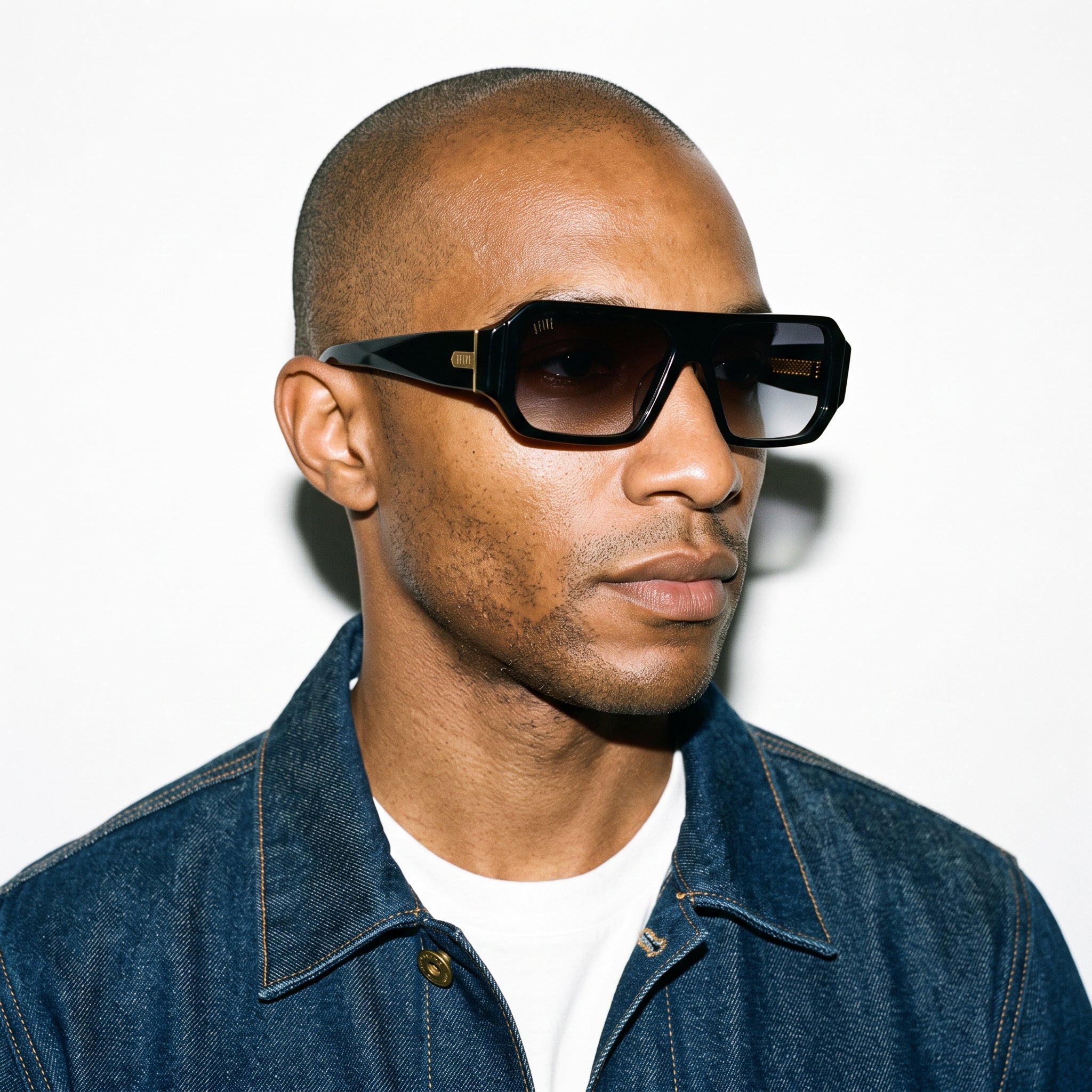 A close-up portrait of a man wearing thick black square sunglasses with dark lenses and a blue denim jacket over a white t-shirt, set against a plain white background. [g:black-gradient s:all o:3]