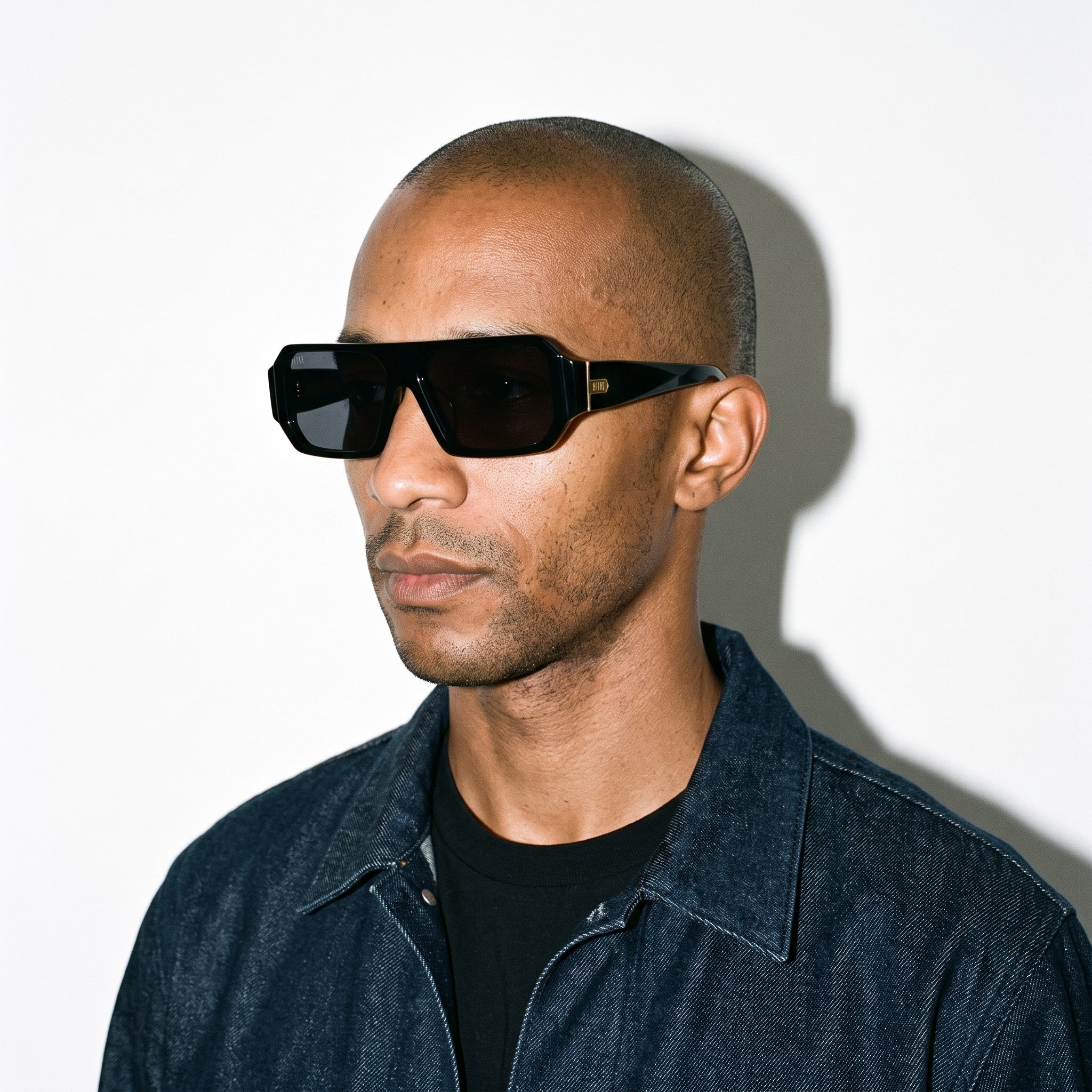 A man with a shaved head wearing black sunglasses and a black denim jacket over a black t-shirt, standing against a white wall with a hard shadow. [g:black s:all o:3]