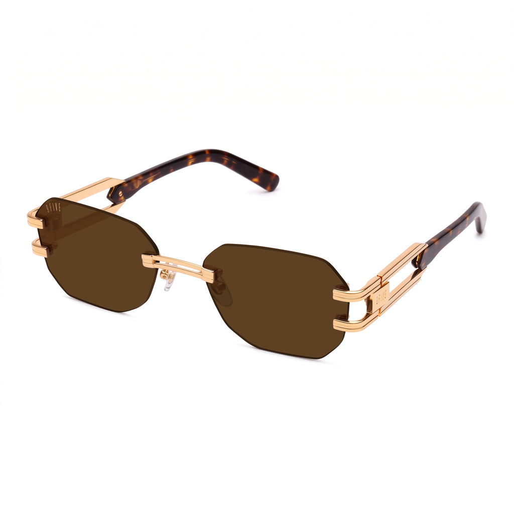 Rimless sunglasses with dark brown lenses, gold metal frames, and tortoiseshell temples, shown on a plain white background. [g:brown s:all o:2]