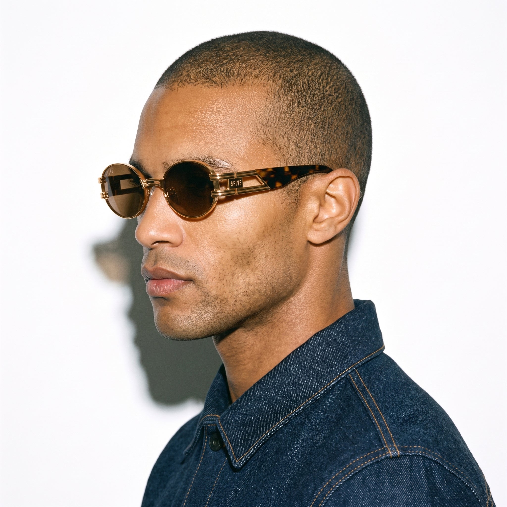 A man in profile wearing round tortoiseshell sunglasses with dark brown lenses and a dark blue denim shirt, set against a plain white background.
[g:brown s:all o:3]