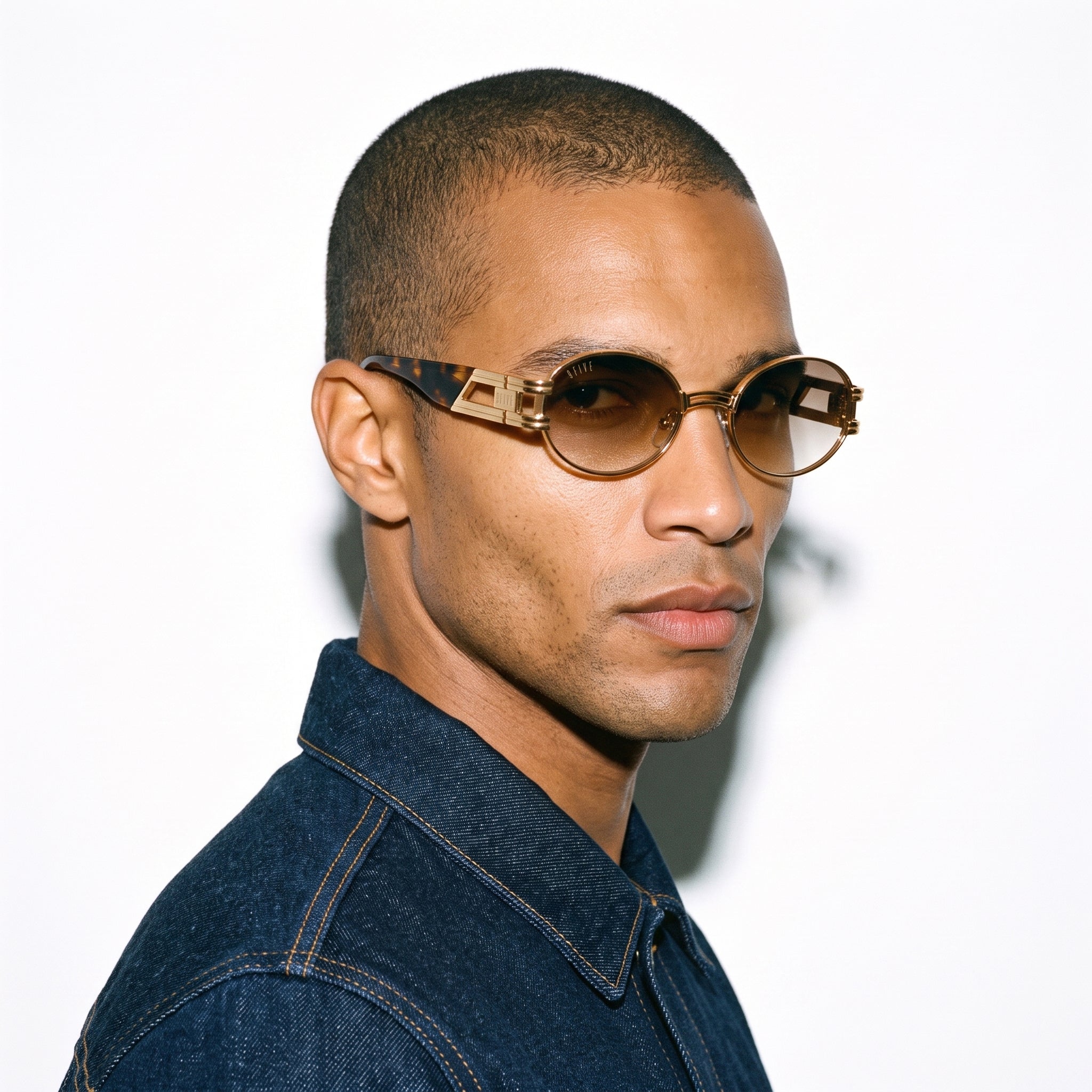 A man in profile wearing round sunglasses with gold metal frames, brown tinted lenses, and geometric tortoiseshell temples, paired with a dark denim shirt against a white background.
[g:brown-gradient s:all o:3]